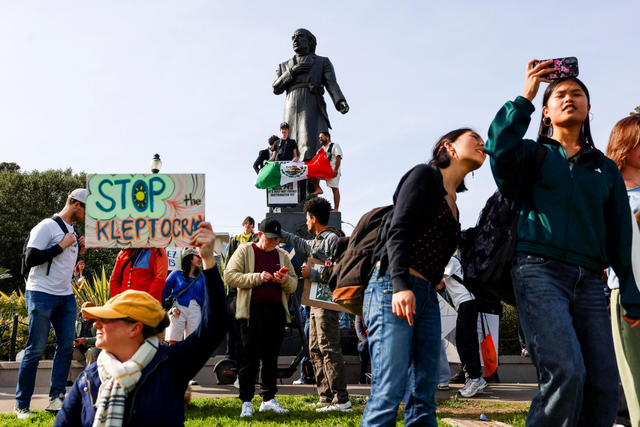 People gather near a statue with some holding signs, including one reading “STOP the KLEPTOCRACY,” while others take photos and interact.